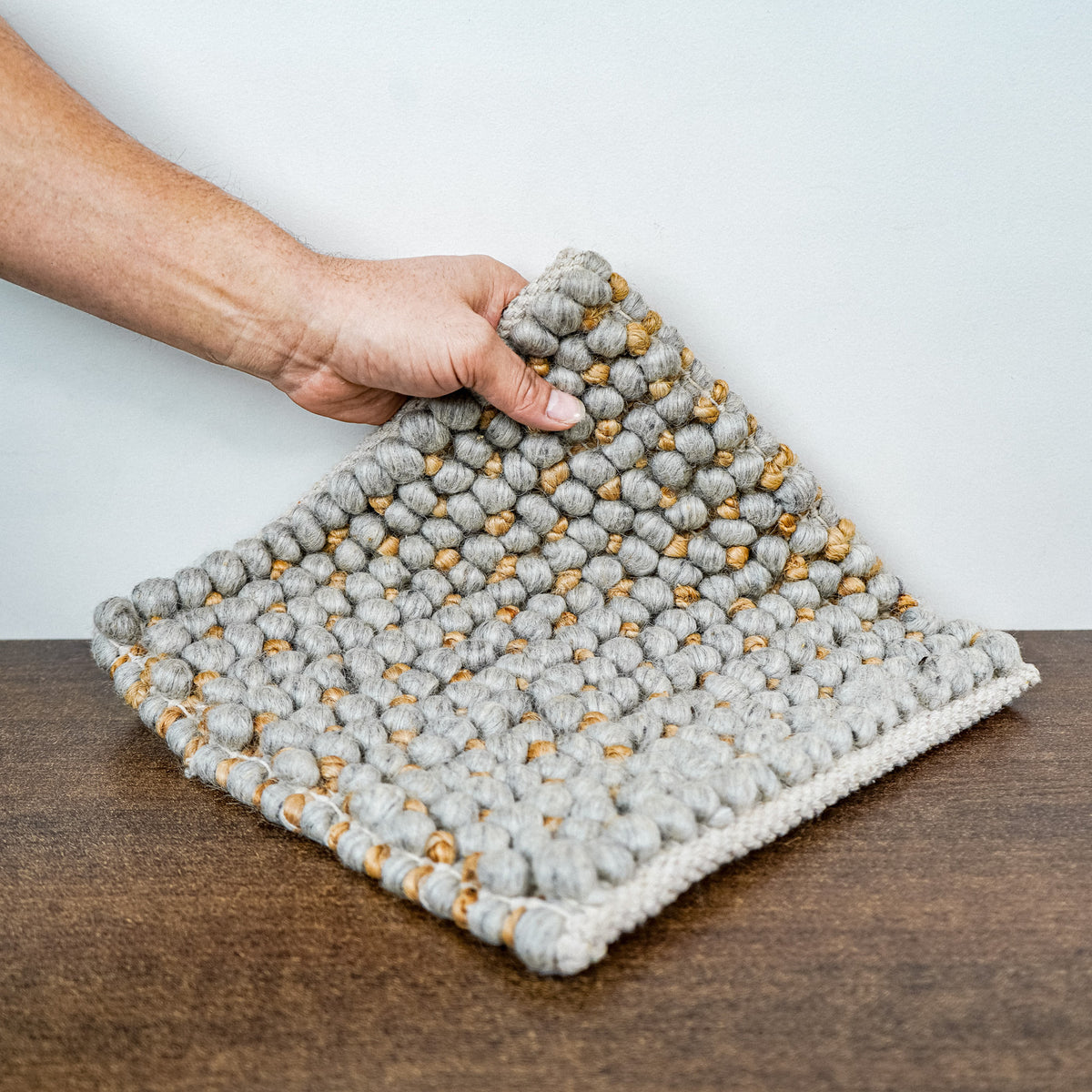 Loops With Jute — gray wool rug sample lifted at one corner by a hand above a brown surface