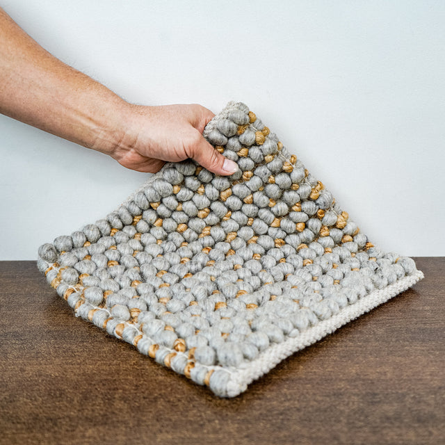 Loops With Jute — gray wool rug sample lifted at one corner by a hand above a brown surface