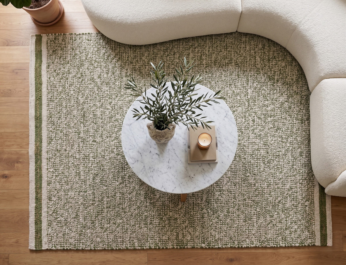 Overhead view of a rectangular green wool rug with striped ends, a round marble table and curved cream sectional.