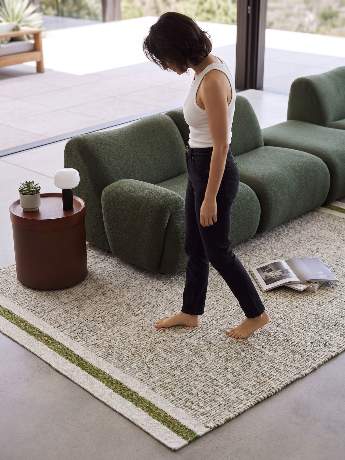 Person walking barefoot across a green wool rug beside a dark green sofa, with the striped end in the foreground.