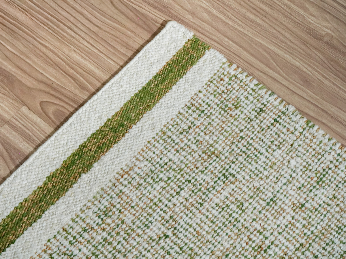 Angled close-up of a green wool rug corner showing the end stripe and green, ivory and tan woven texture.
