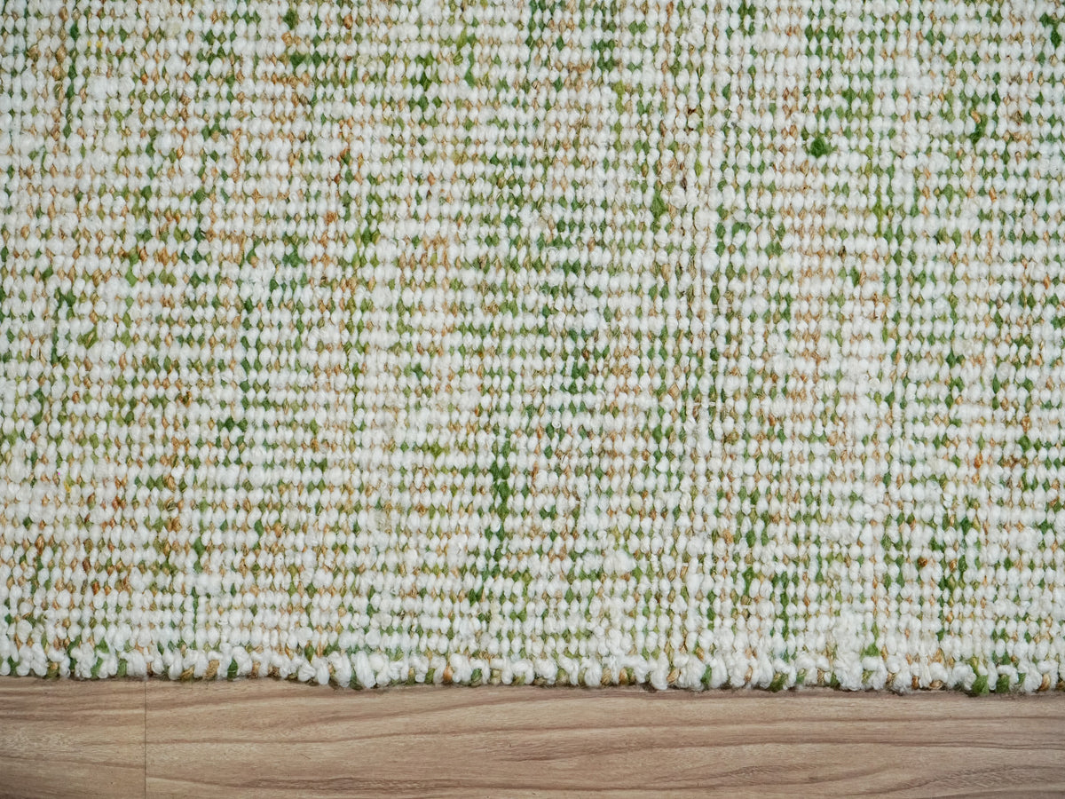 Top-down detail of the green wool rug's closely spaced ivory yarns, green accents and edge against a wood floor.