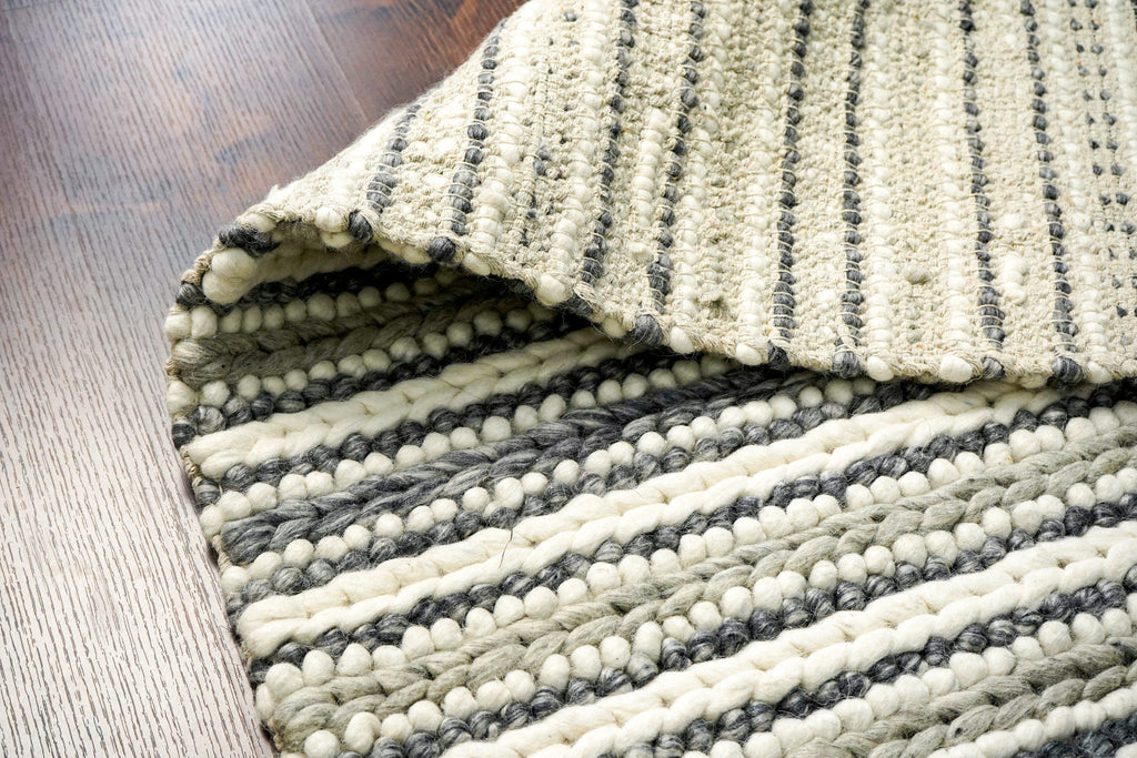 May include: A close-up of a woven rug with a striped pattern. The rug is made of a combination of white, gray, and beige yarn. The rug has a textured surface with small, round pom-poms throughout.