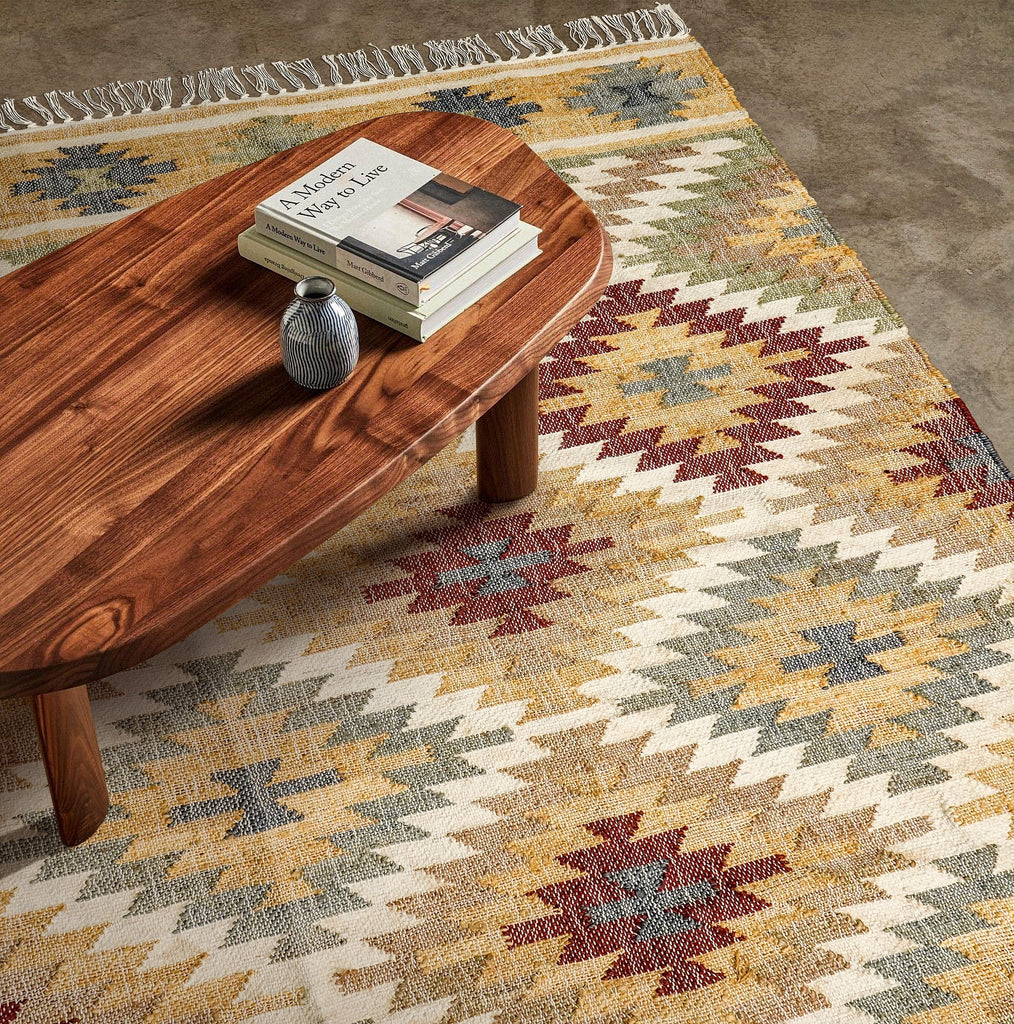 May include: A wooden coffee table with a rounded oval top and four legs. The table is sitting on a rug with a geometric pattern in shades of brown, red, and blue.
